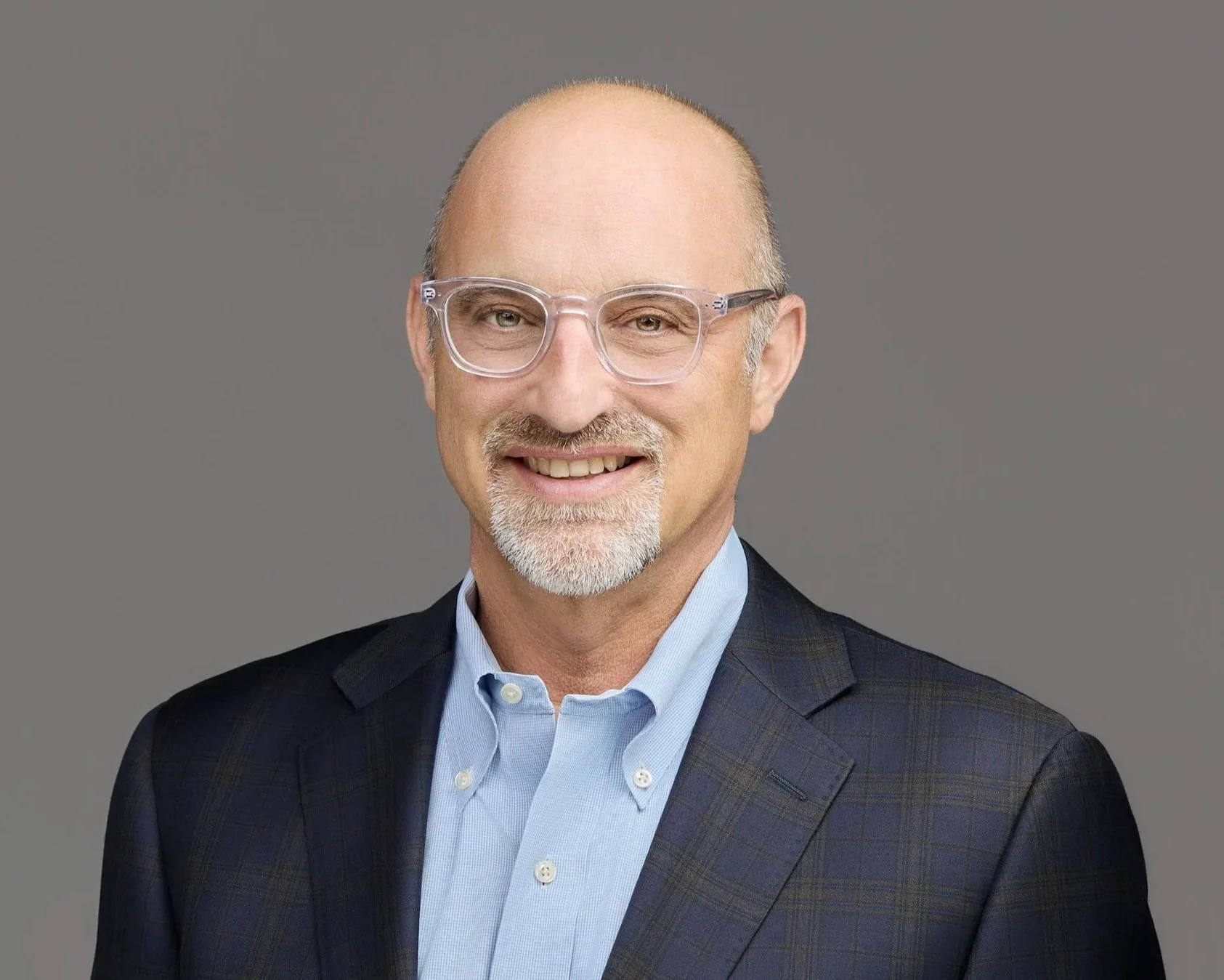 Middle-aged man with a bald head, wearing clear glasses, a light blue dress shirt, and a dark plaid blazer, smiling against a neutral gray background.