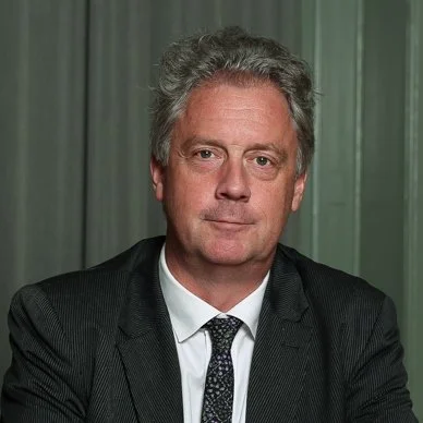 Portrait of a middle-aged man with gray hair, wearing a dark suit, white shirt, and patterned tie, standing against a green curtain background.