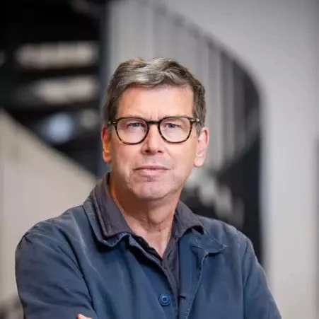 A middle-aged man with gray hair and glasses, standing indoors with a neutral expression, wearing a dark jacket over a shirt.