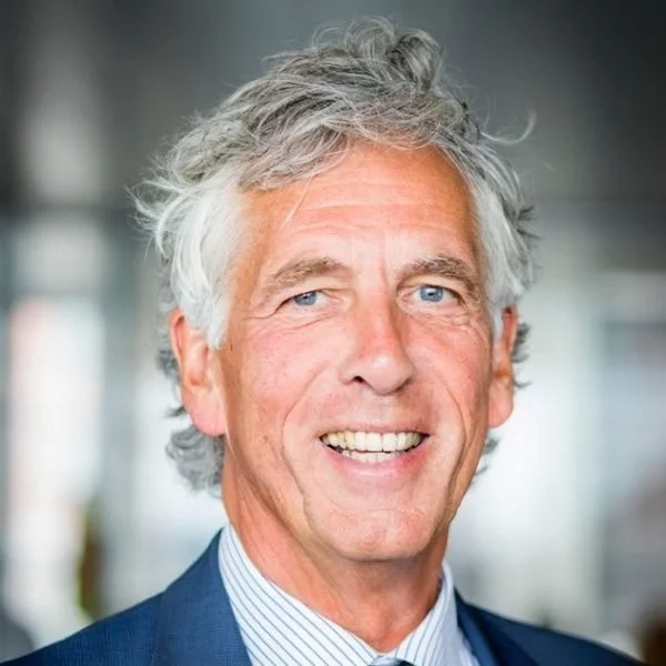 Close-up photo of a smiling middle-aged man with gray hair, blue eyes, wearing a dark suit and a light striped shirt, in a professional indoor setting.