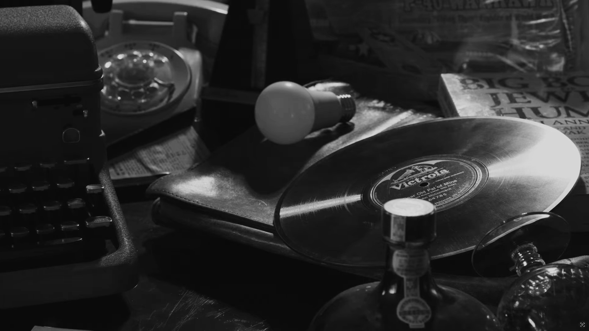 A cluttered tabletop with a vinyl record, a stack of books, a light bulb, a rotary phone, and various small objects in black and white.
