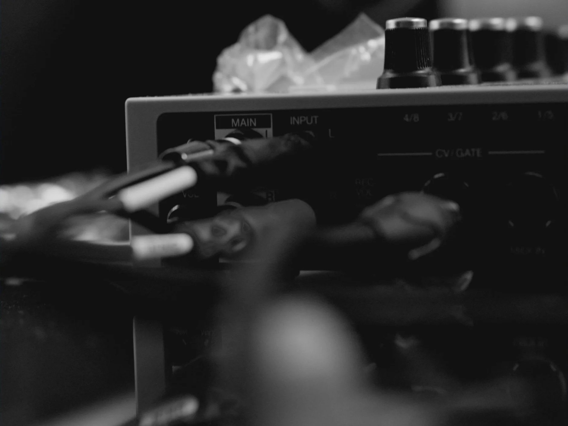 A close-up of a mixing console with various knobs and sliders, some cables connected, and a plastic bag with crumpled paper in the background.