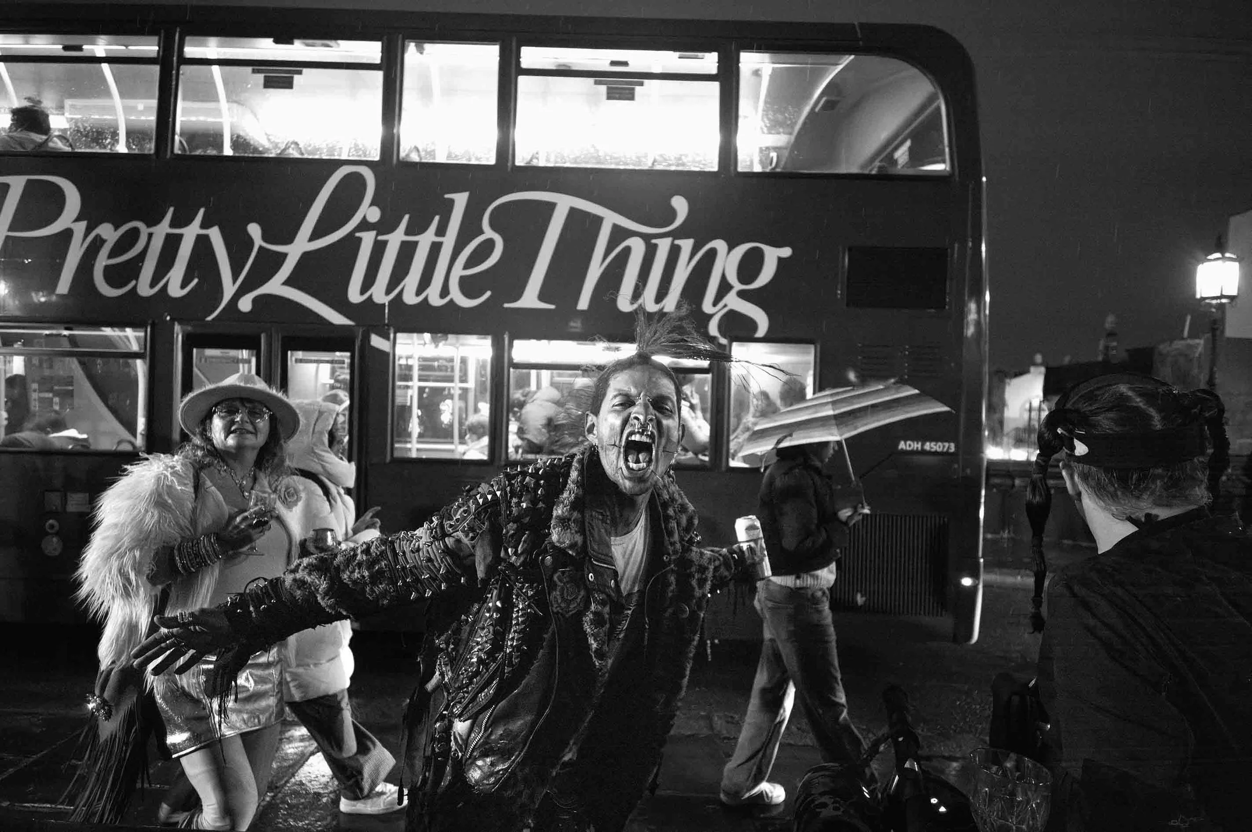 Black and white street photograph of a punk shouting in front of a double-decker bus in Camden Town, London at night