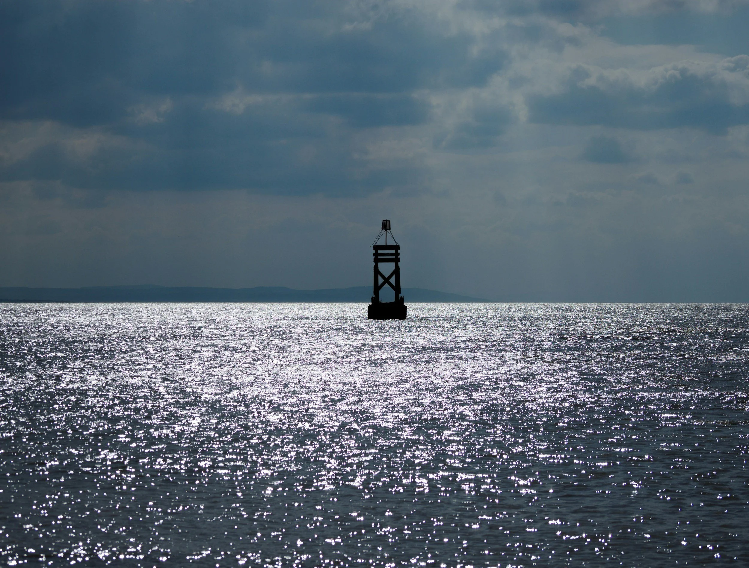 A silhouette of a small lighthouse or buoy in the water, with a distant landmass on the horizon and a cloudy sky overhead.