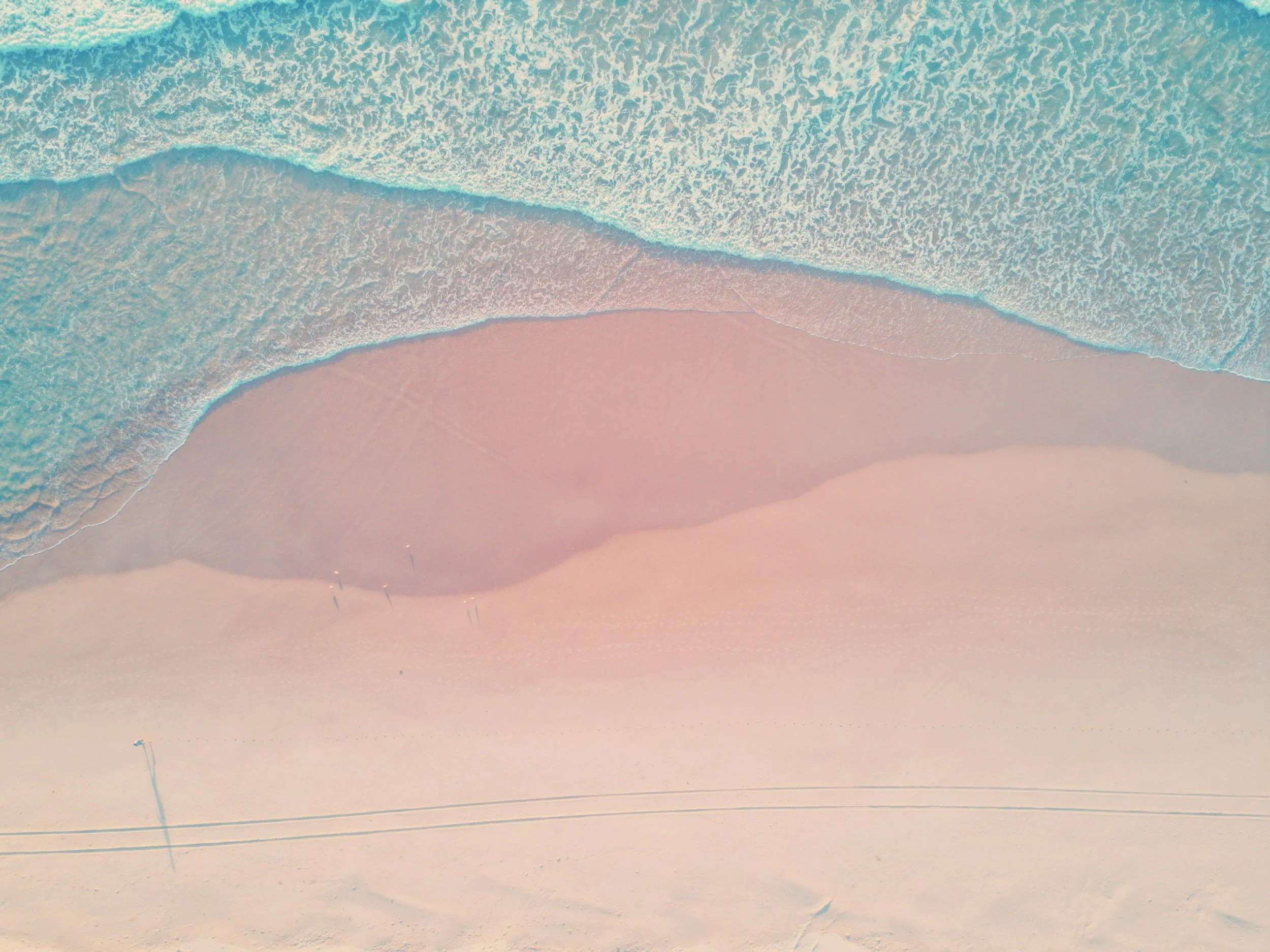 Aerial view of a beach with pink sand, turquoise ocean waves, and a few people walking along the shoreline.