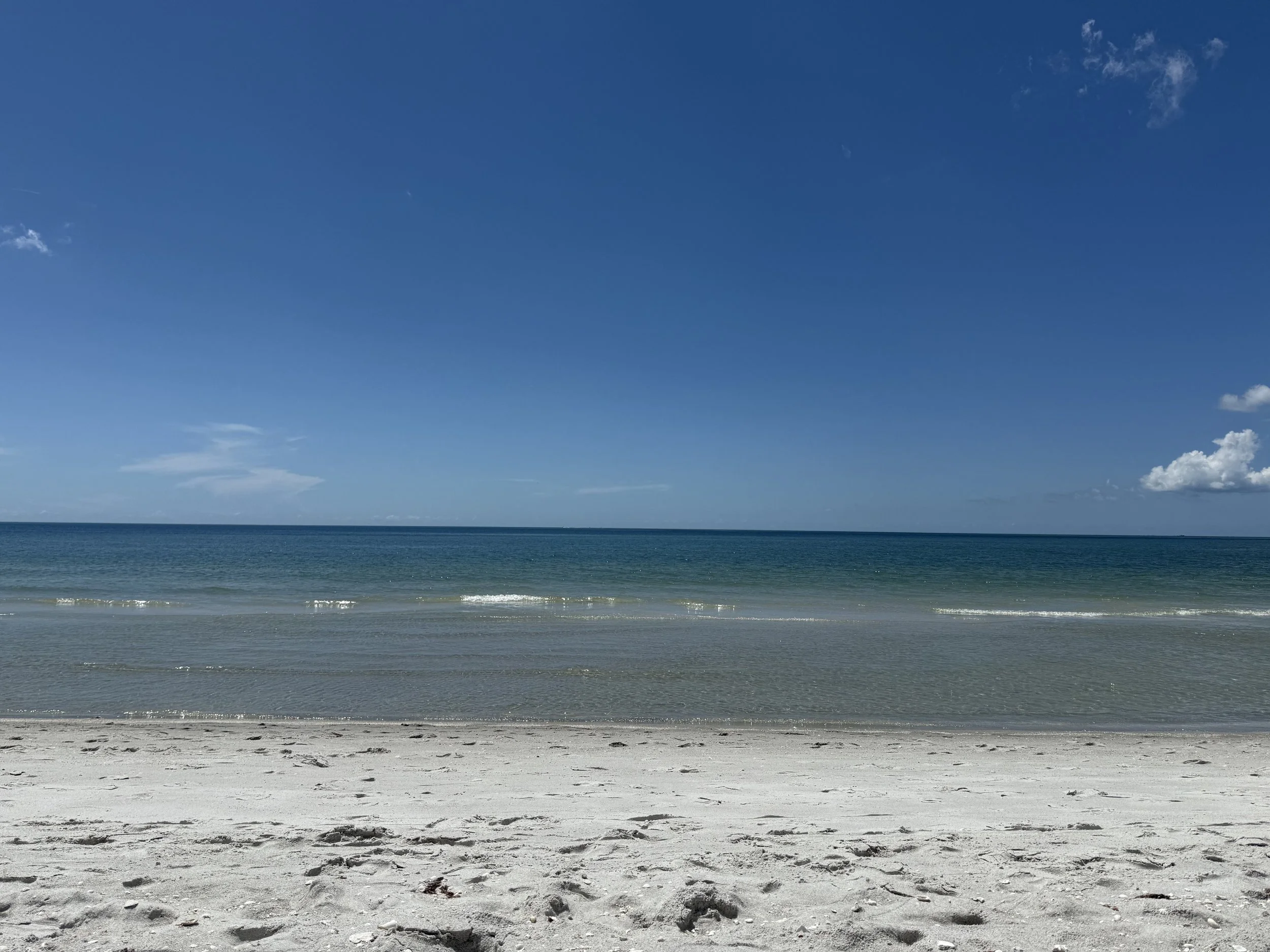 Bonita Springs, FL, A serene beach scene with white sandy shore, calm ocean waters, and a clear blue sky with a few clouds.
