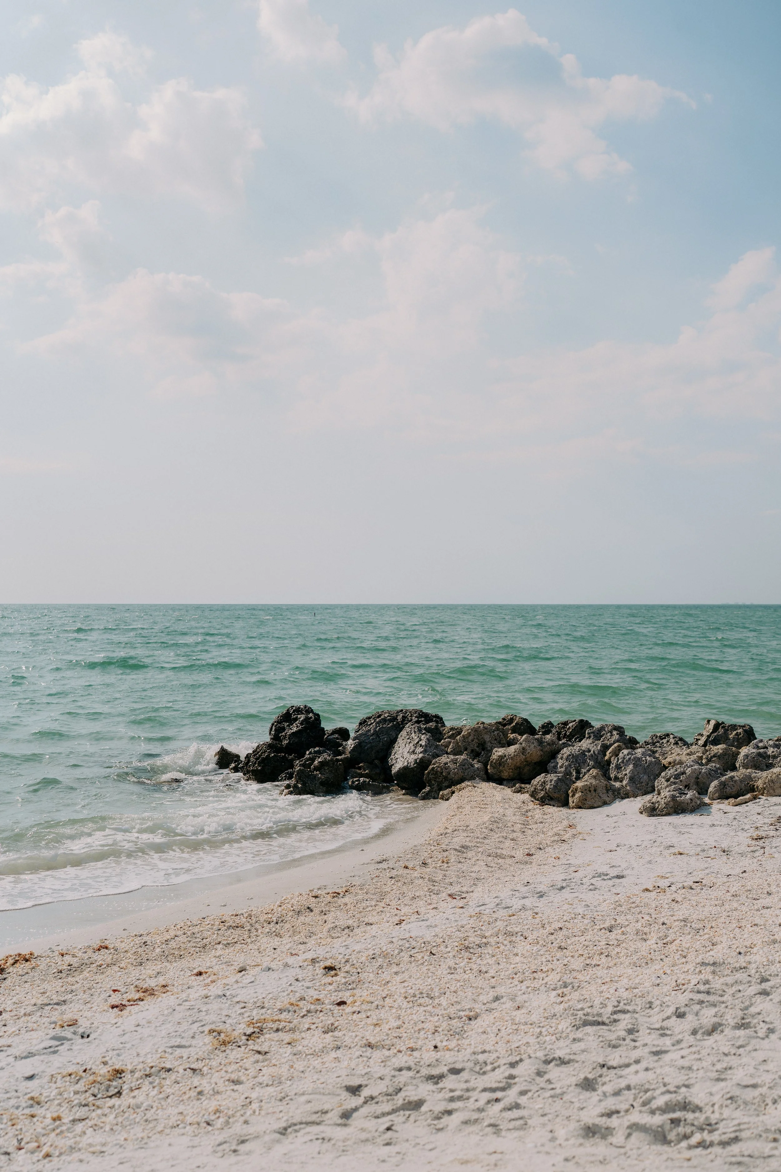 A sandy beach with rocks near the shoreline, calm ocean water, and a partly cloudy sky.
