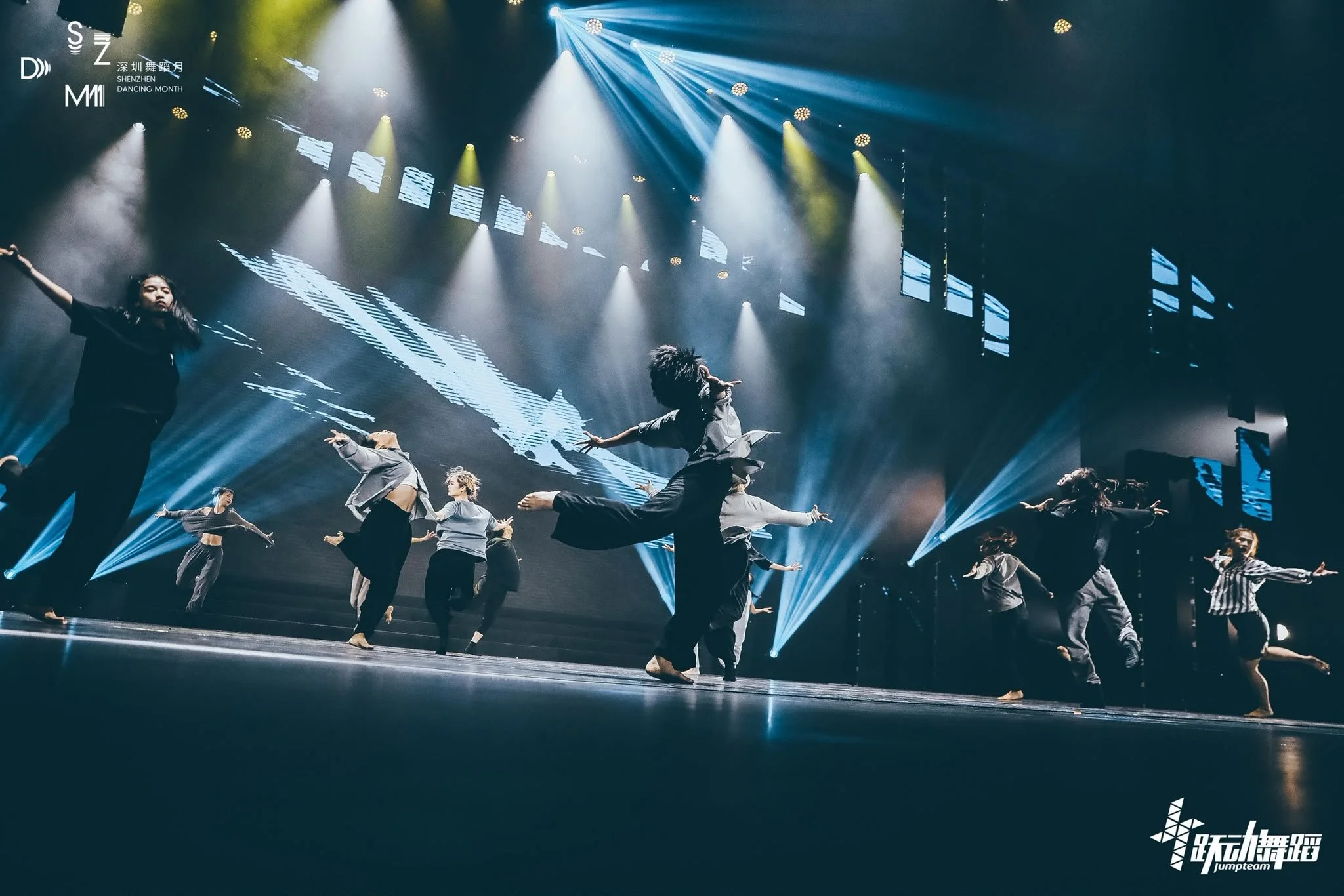 Dance performance on stage with multiple dancers dressed in casual attire, illuminated by blue and yellow spotlights, with digital screens displaying abstract visuals in the background.