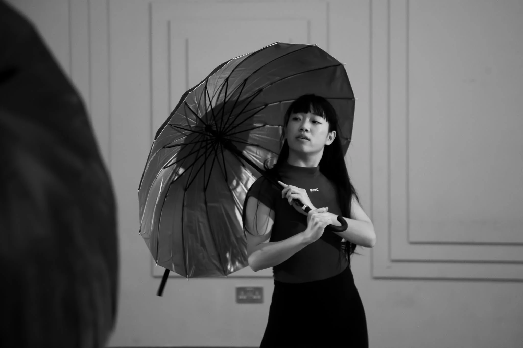 A woman holds an umbrella and looks carefully at something or someone outside the frame in a room with plain walls.