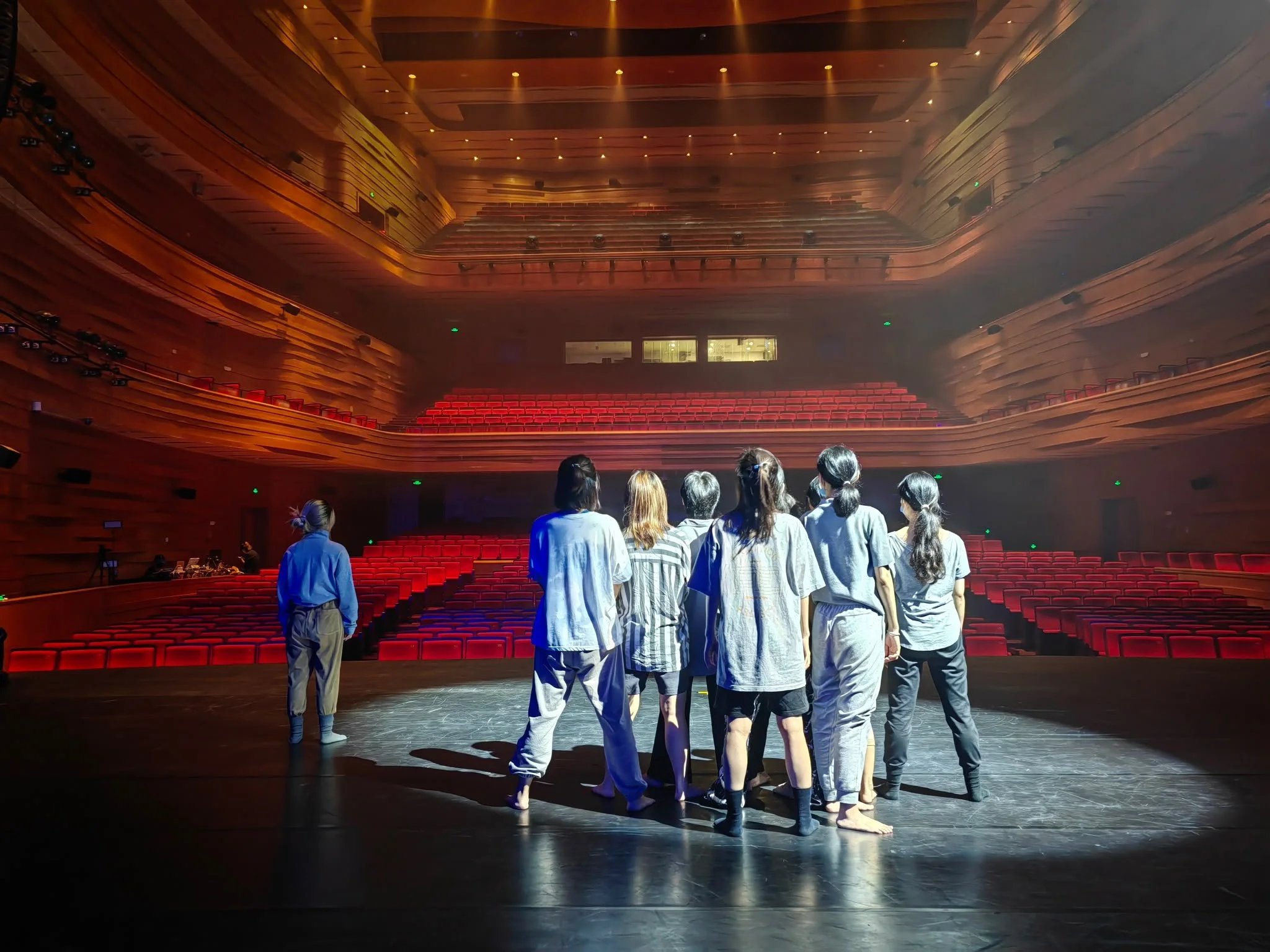 A group of people in casual clothing standing barefoot on a stage in a large auditorium with red seating and wooden interior, preparing for a performance.