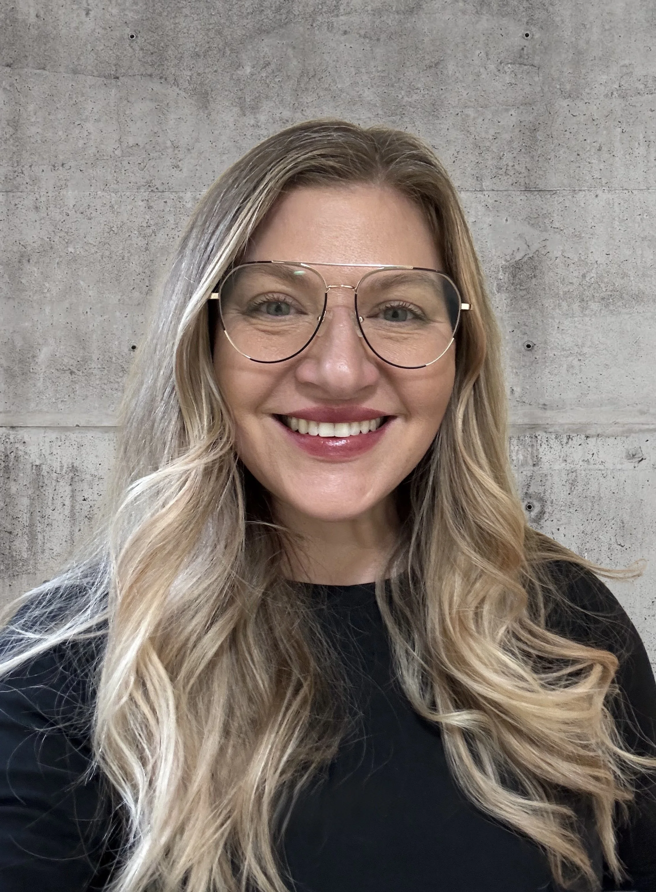 A woman with long, wavy blonde hair, glasses, and a black shirt smiling in front of a gray concrete wall.
