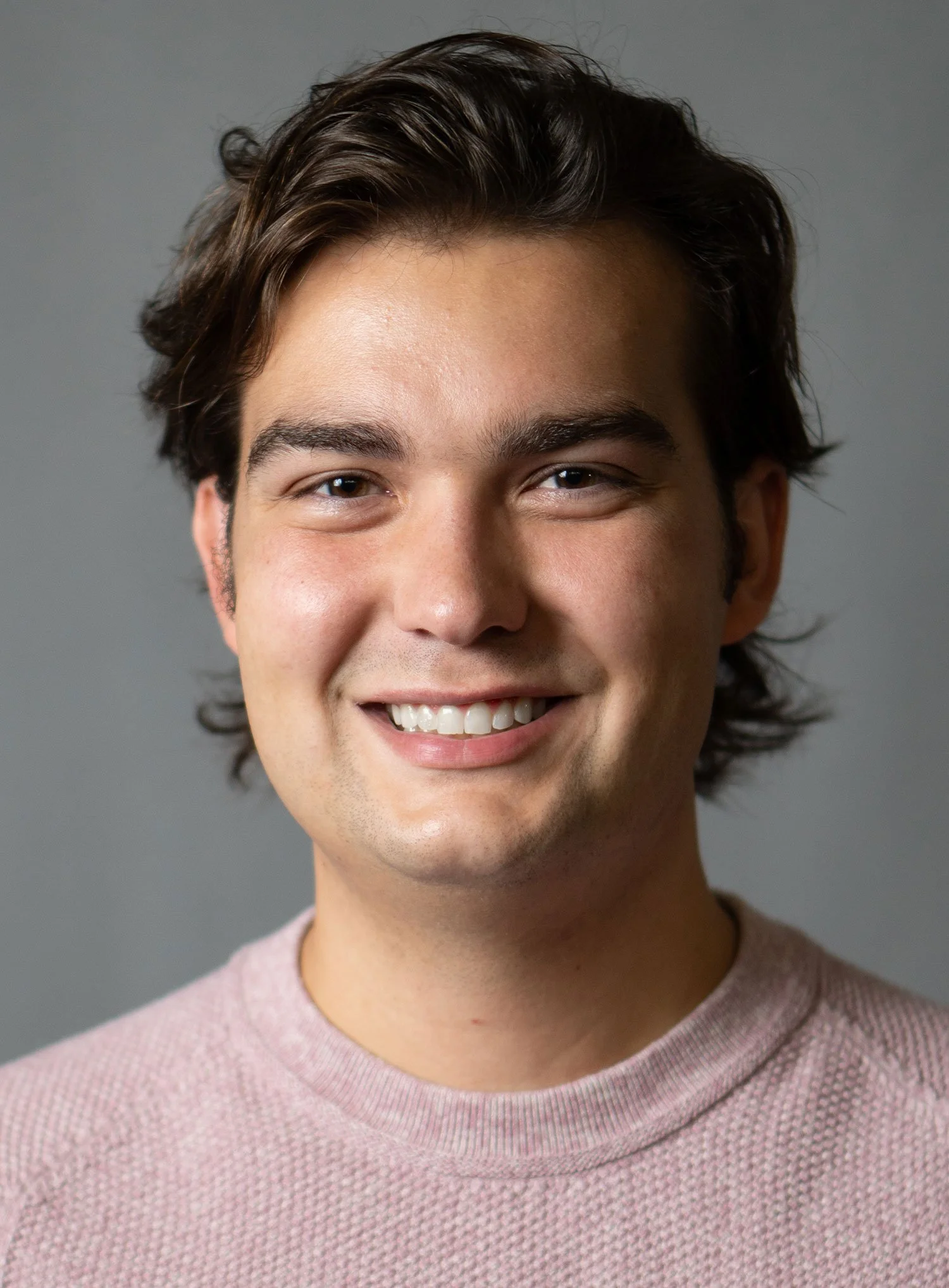 A young man with dark wavy hair and a broad smile, wearing a light pink textured shirt, standing against a neutral gray background.
