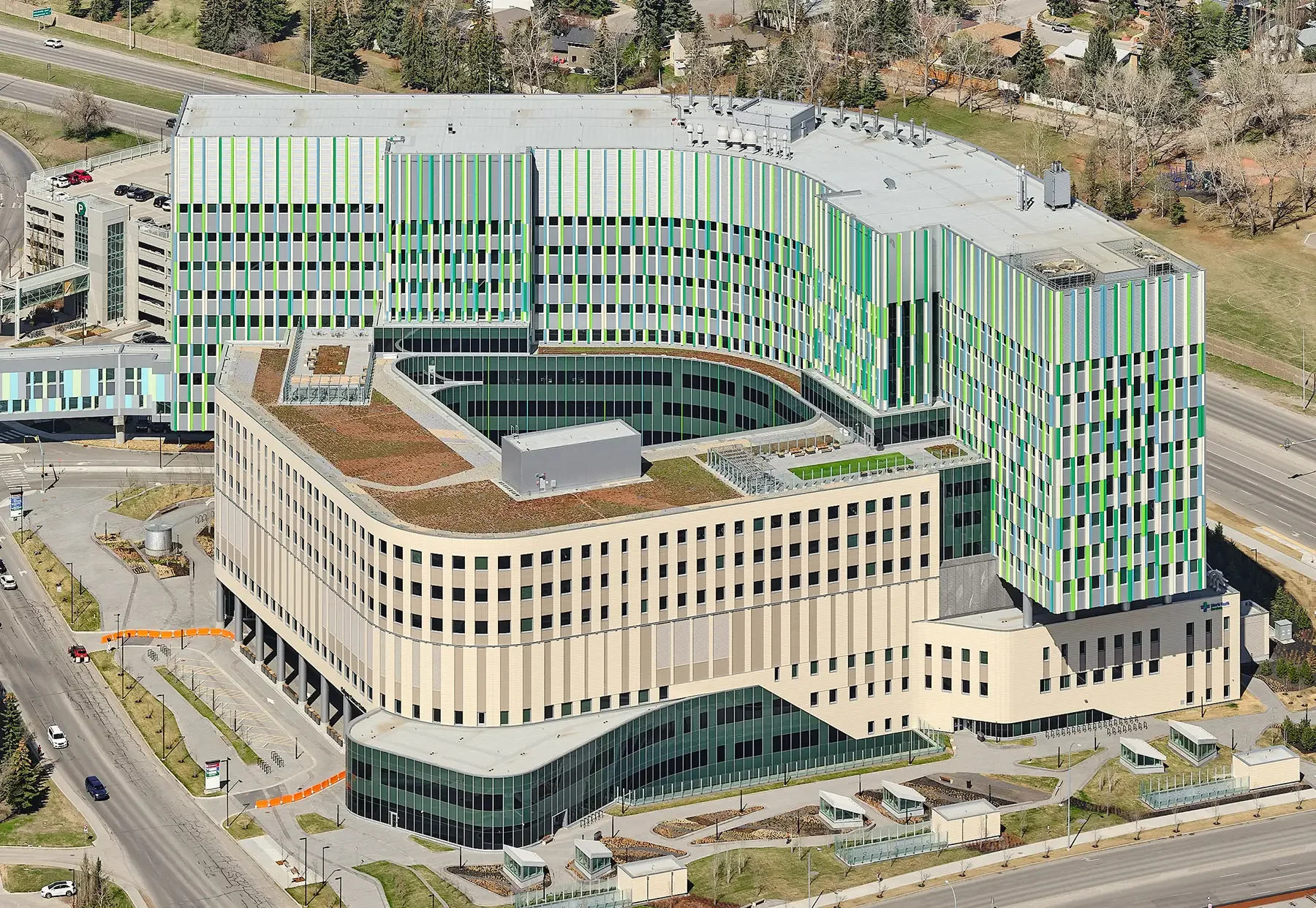 Aerial view of a modern hospital building with a colorful, patterned facade, green roofing, and surrounding roads and green spaces.