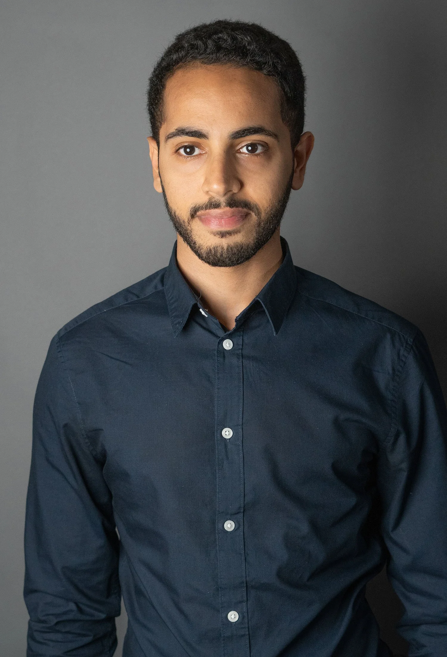Portrait of a young man with a beard and short curly hair, wearing a dark blue button-up shirt, standing against a gray background.