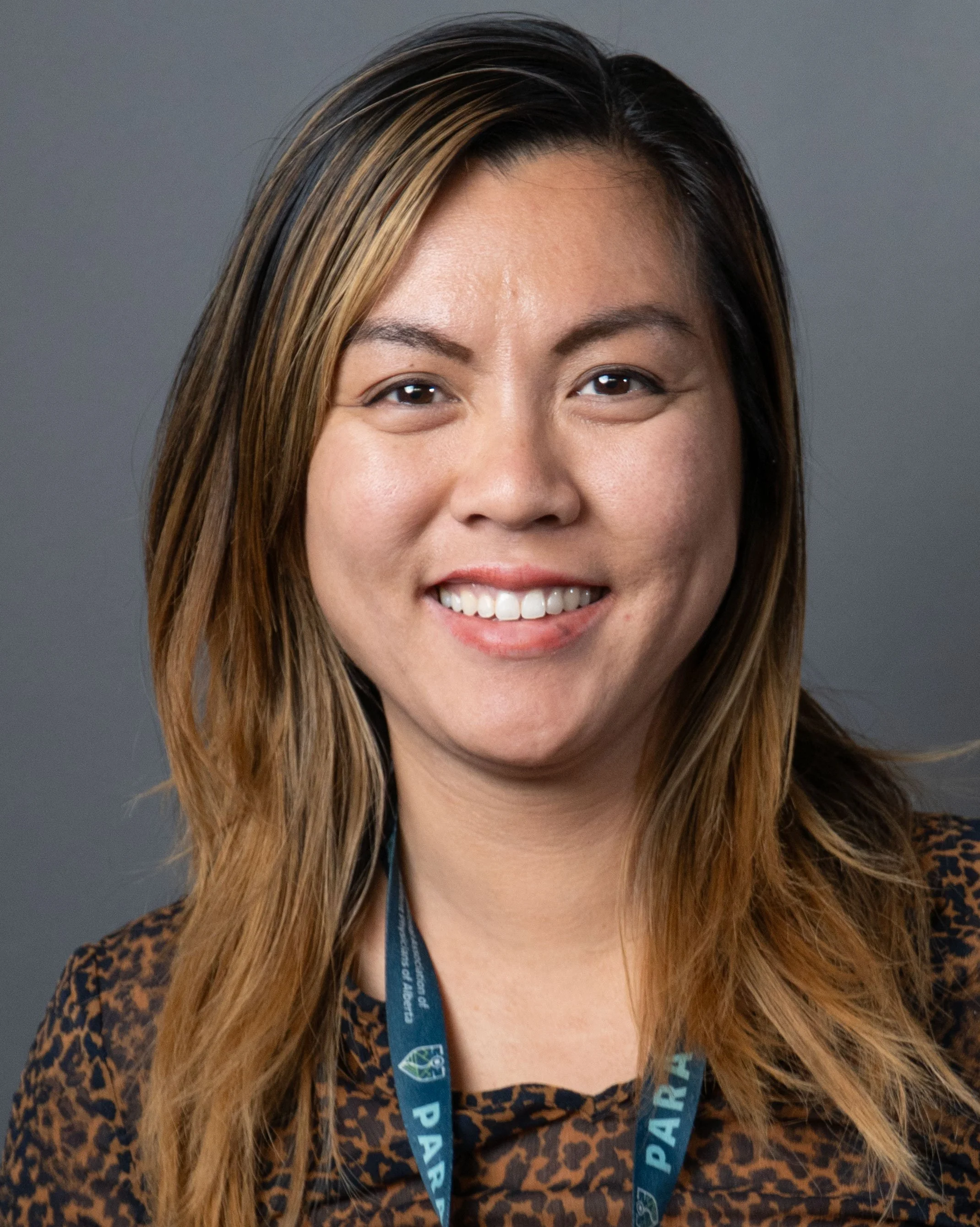 A woman with shoulder-length hair, wearing a leopard print top and a lanyard, smiling against a gray background.