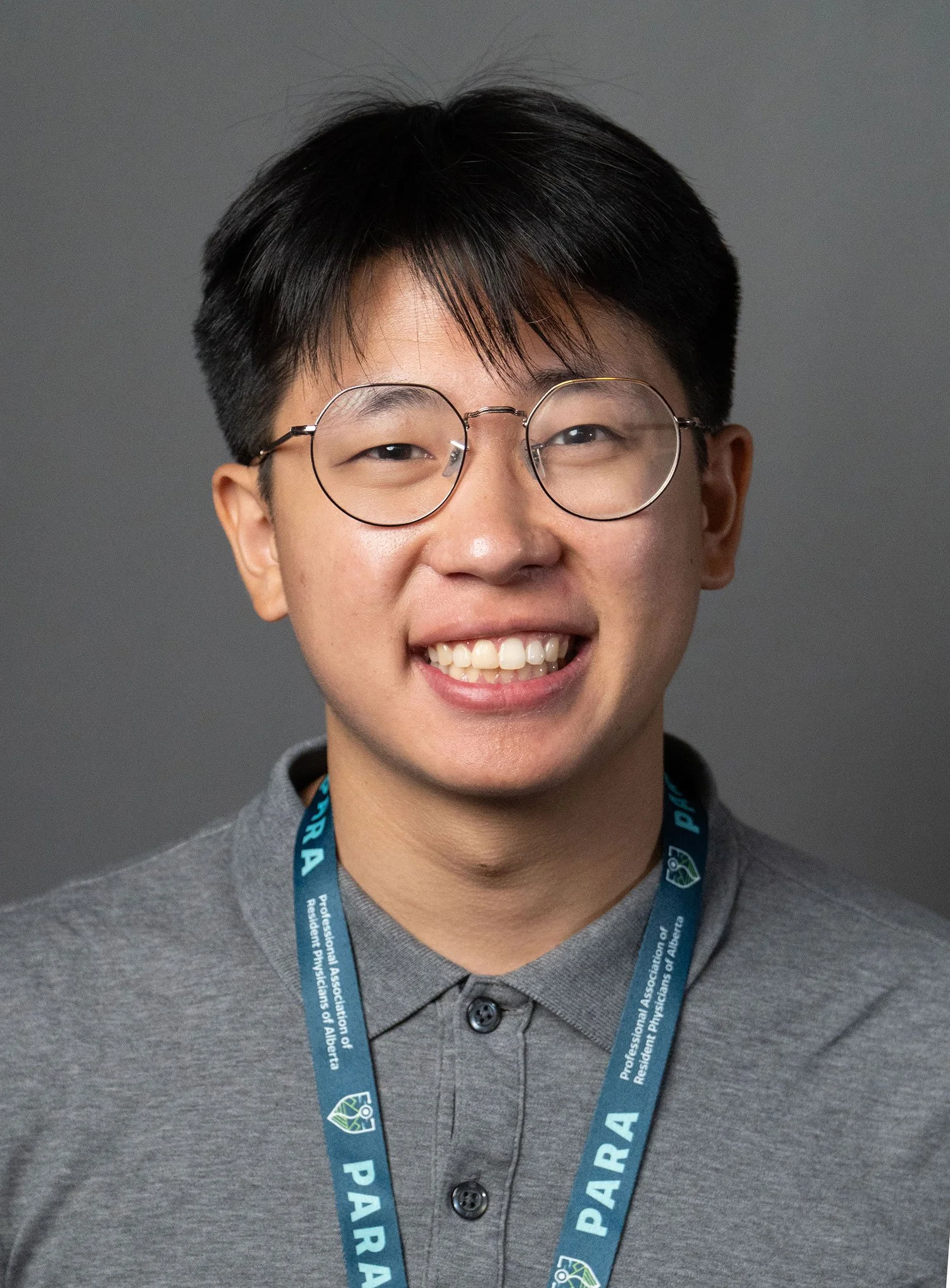 Young man smiling, wearing glasses, a gray button-up shirt, and a lanyard that reads 'PARA' with the full form 'Professional Association of Resident Physicians of Alberta' visible, against a gray background.