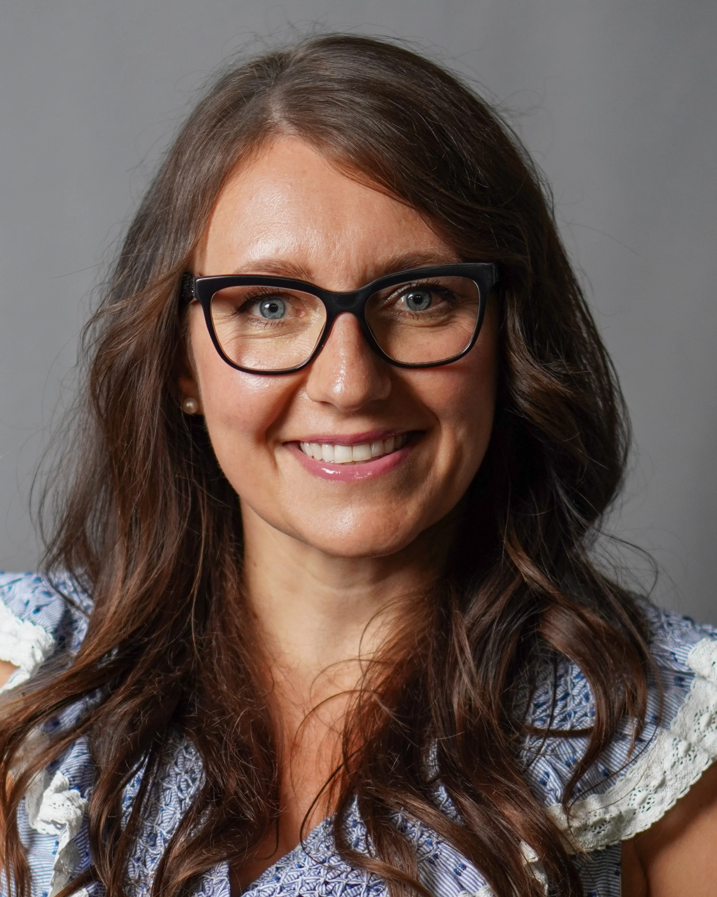 A woman with long brown hair, wearing black glasses and a blue patterned top, smiling at the camera against a plain gray background.