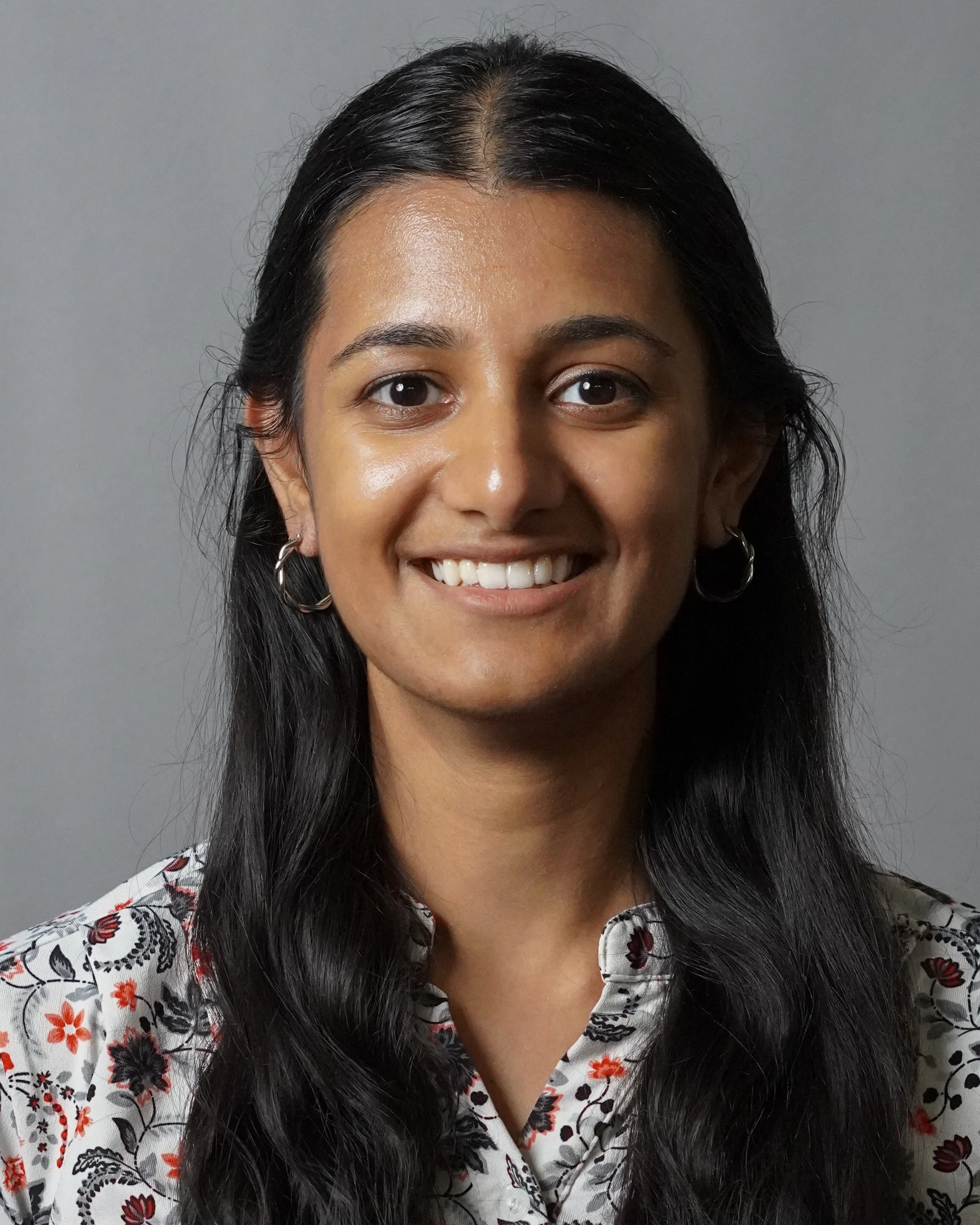 A woman with long black hair, wearing hoop earrings and a floral patterned blouse, smiling against a gray background.