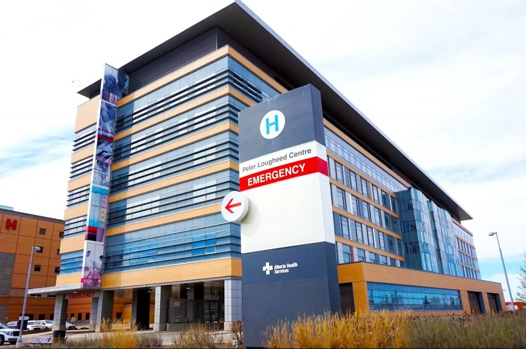 Modern hospital building with a large sign indicating Peter Lougheed Centre Emergency, operated by Alberta Health Services, under a partly cloudy sky.
