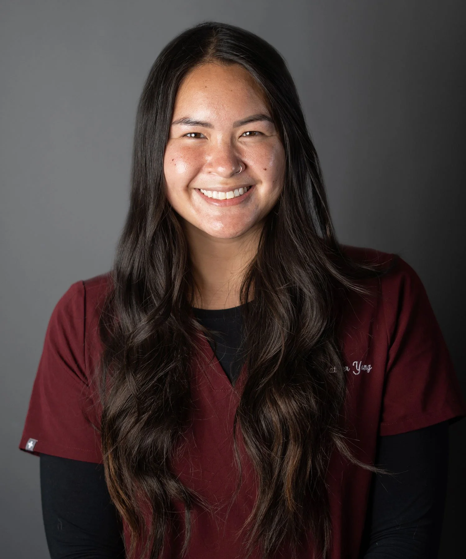 Young woman with long dark hair smiling, wearing a maroon medical uniform against a gray background.
