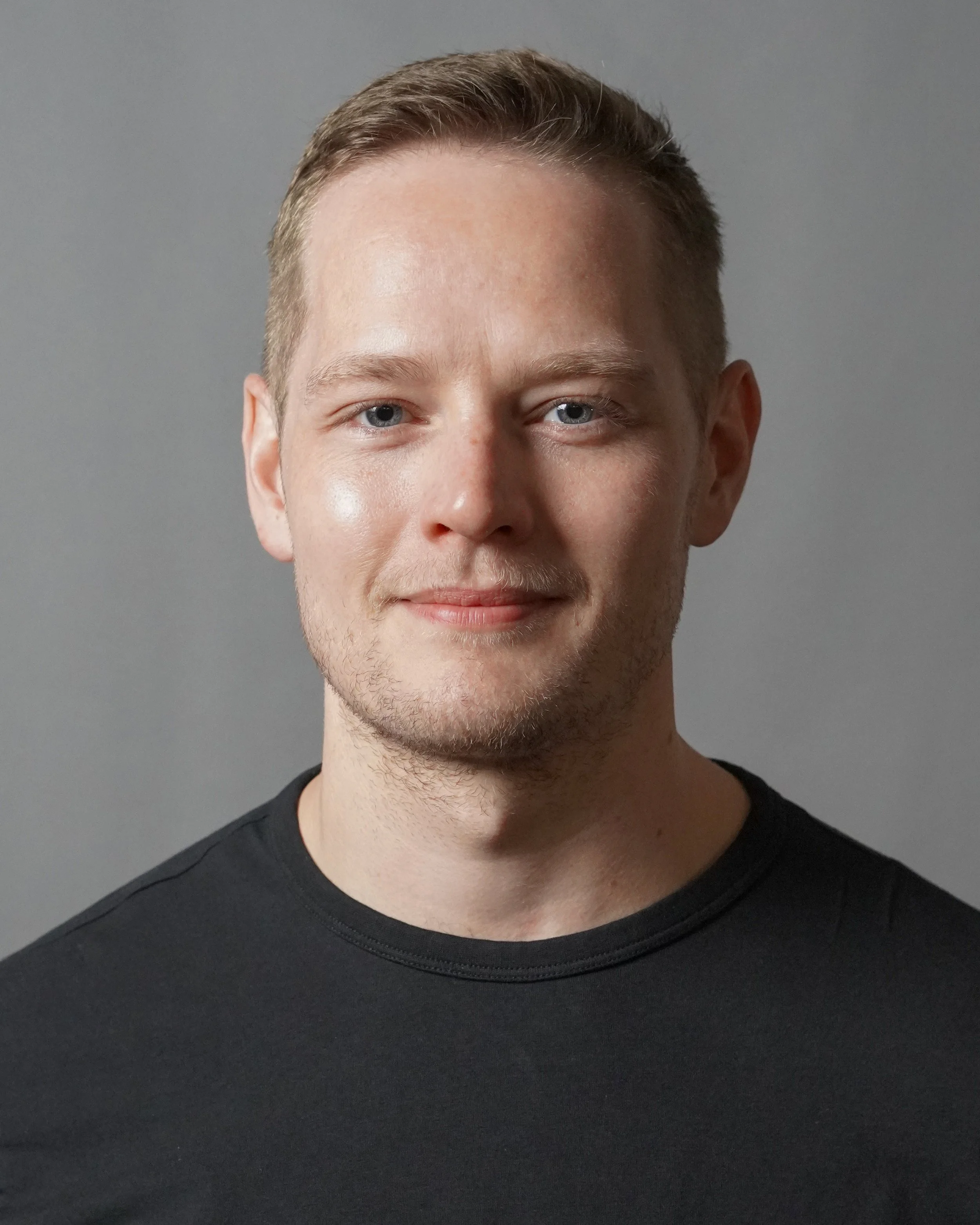 A young man with short, light brown hair and blue eyes, wearing a black shirt, smiling slightly, against a plain gray background.