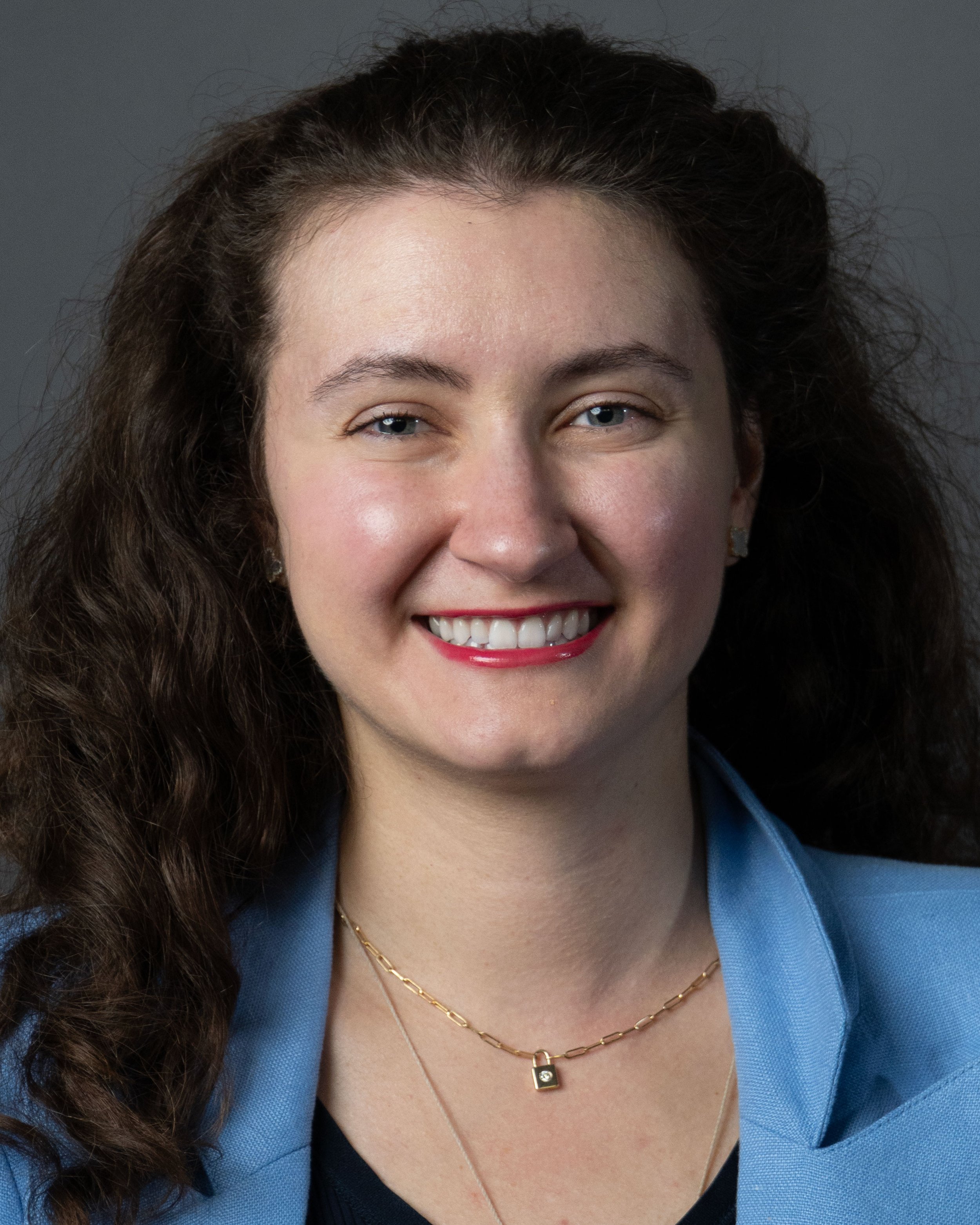 Portrait of a woman with long curly brown hair, light skin, blue eyes, wearing a light blue blazer, gold jewelry, and red lipstick, smiling against a gray background.