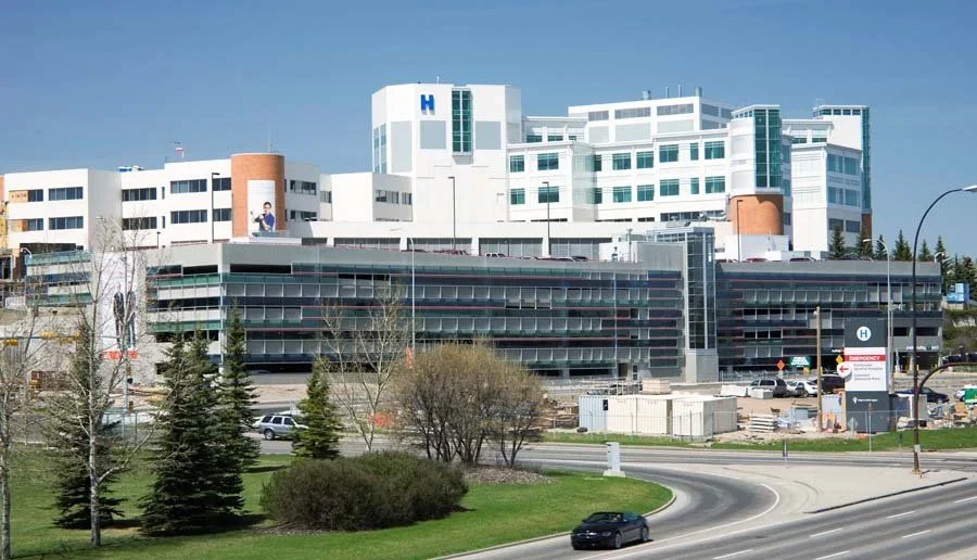 Large modern hospital building with white and gray exterior, multiple windows, and a prominent blue letter "H" sign, situated near a curved road with some trees and greenery in the foreground.