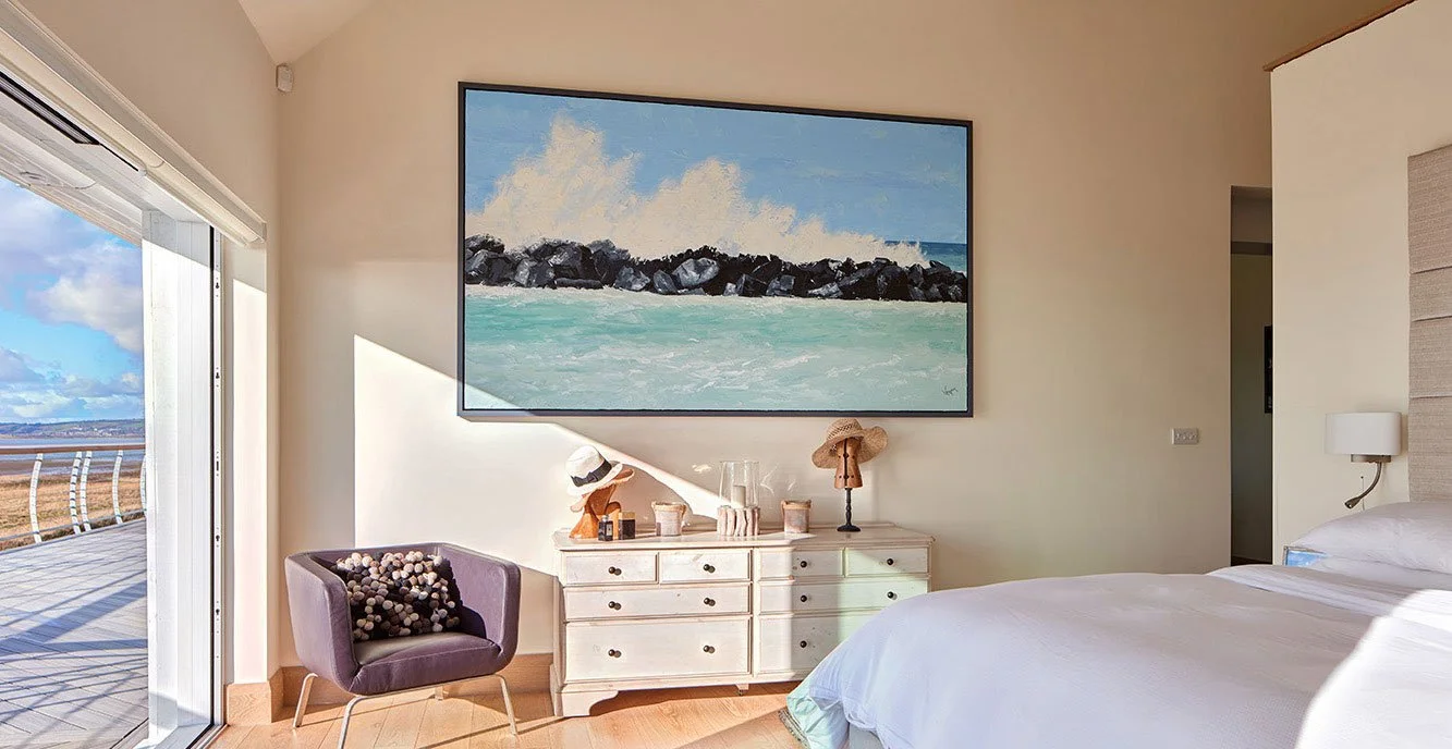 A bright hotel room with a balcony view of a blue sky and open landscape, a white bed, a purple armchair with a floral pillow, a white dresser with decorative items, and a large seascape painting on the wall.