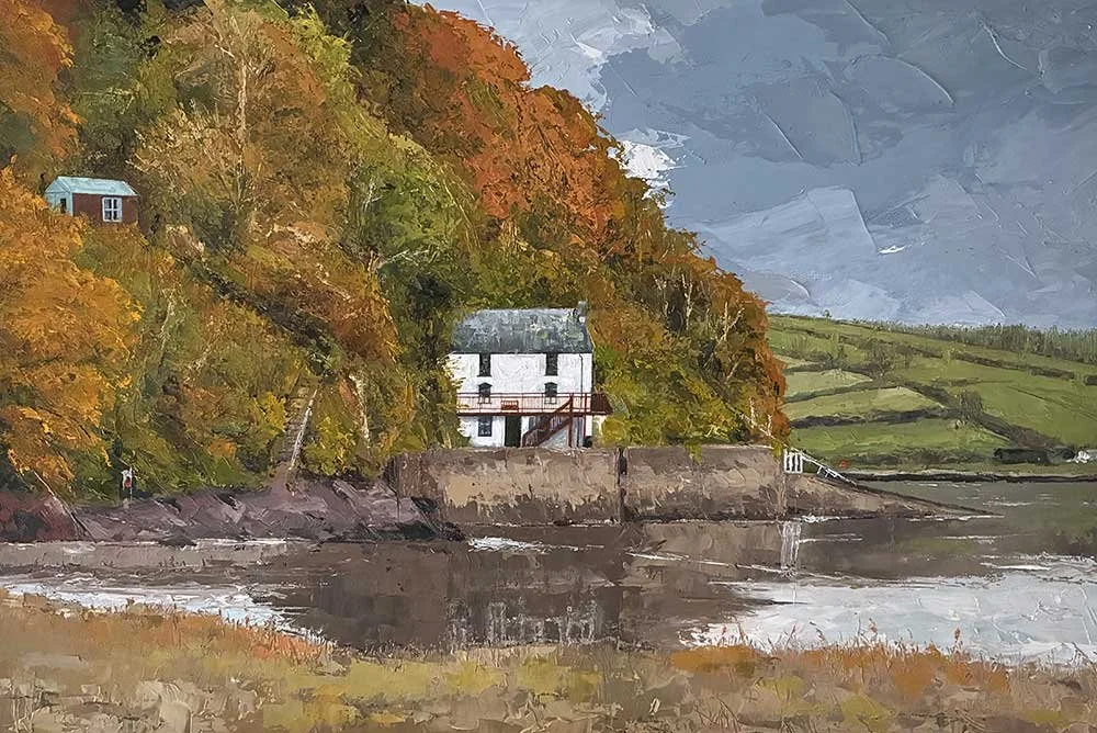 A painting of a riverside scene with a white house at the base of a hill covered in orange and green autumn trees, with stormy skies overhead and another small house on the hillside. DYLAN THOMAS BOATHOUSE