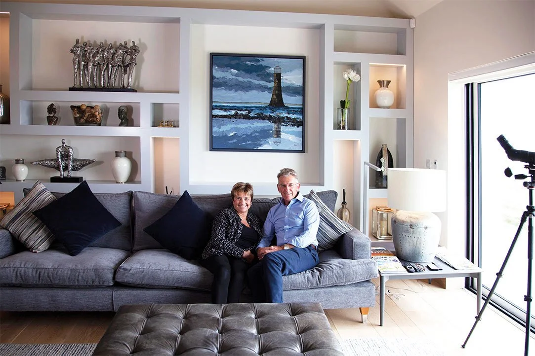 A living room with a gray sofa, two people sitting on it, a large painting of a lighthouse on the wall, decorative shelves with vases and sculptures, a side table with a lamp, and a telescope near a sliding glass door.