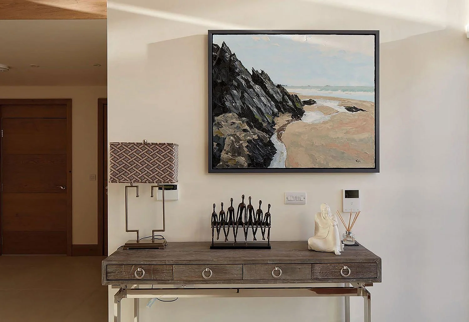 A wooden console table with decorative items, a table lamp, a sculpture, a figurine group, and a reed diffuser, mounted on a white wall with a large framed seascape painting above.