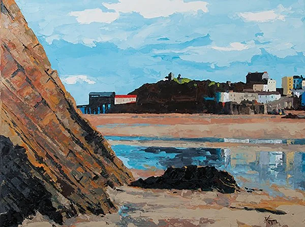 Ocean shoreline with a large rock, a sandy beach, and a distant town with buildings and greenery against a partly cloudy sky.