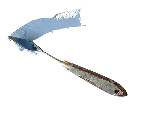 A metal putty knife with a wooden handle applying blue painter's putty or modeling compound on a white surface.