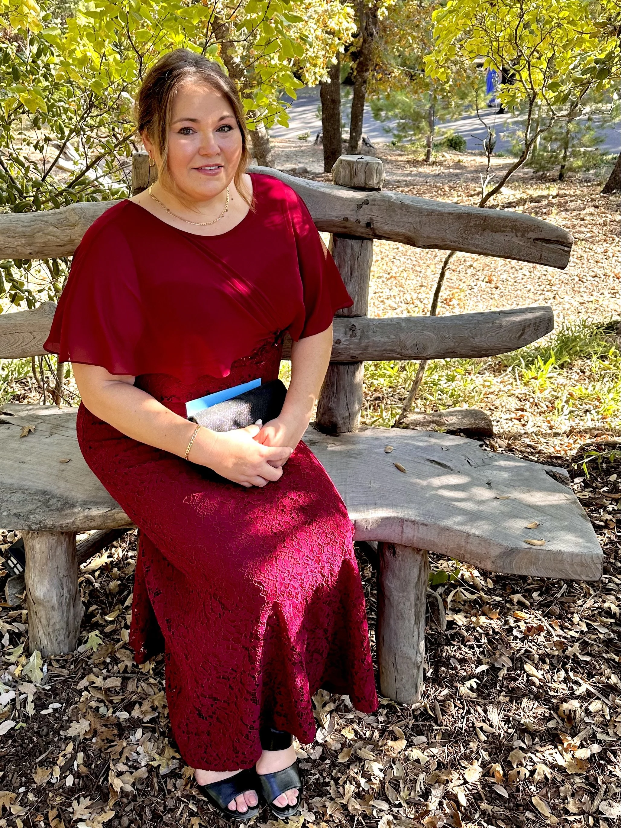 A woman in a red dress sitting on a wooden bench outdoors surrounded by trees and fallen leaves.