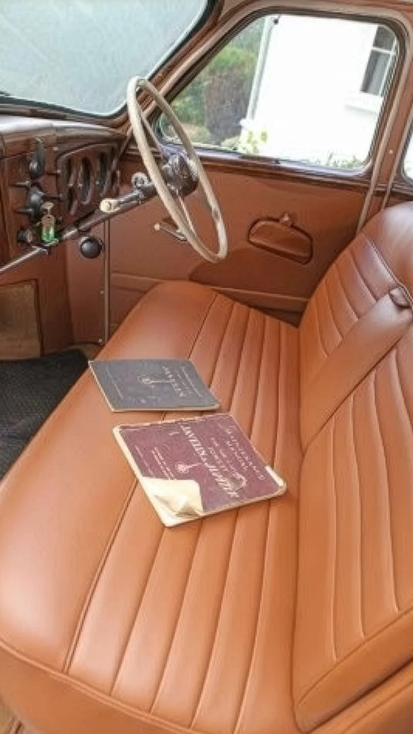 Interior of a vintage car with a tan leather bench seat, a brown dashboard, and a steering wheel. Two books rest on the seat.