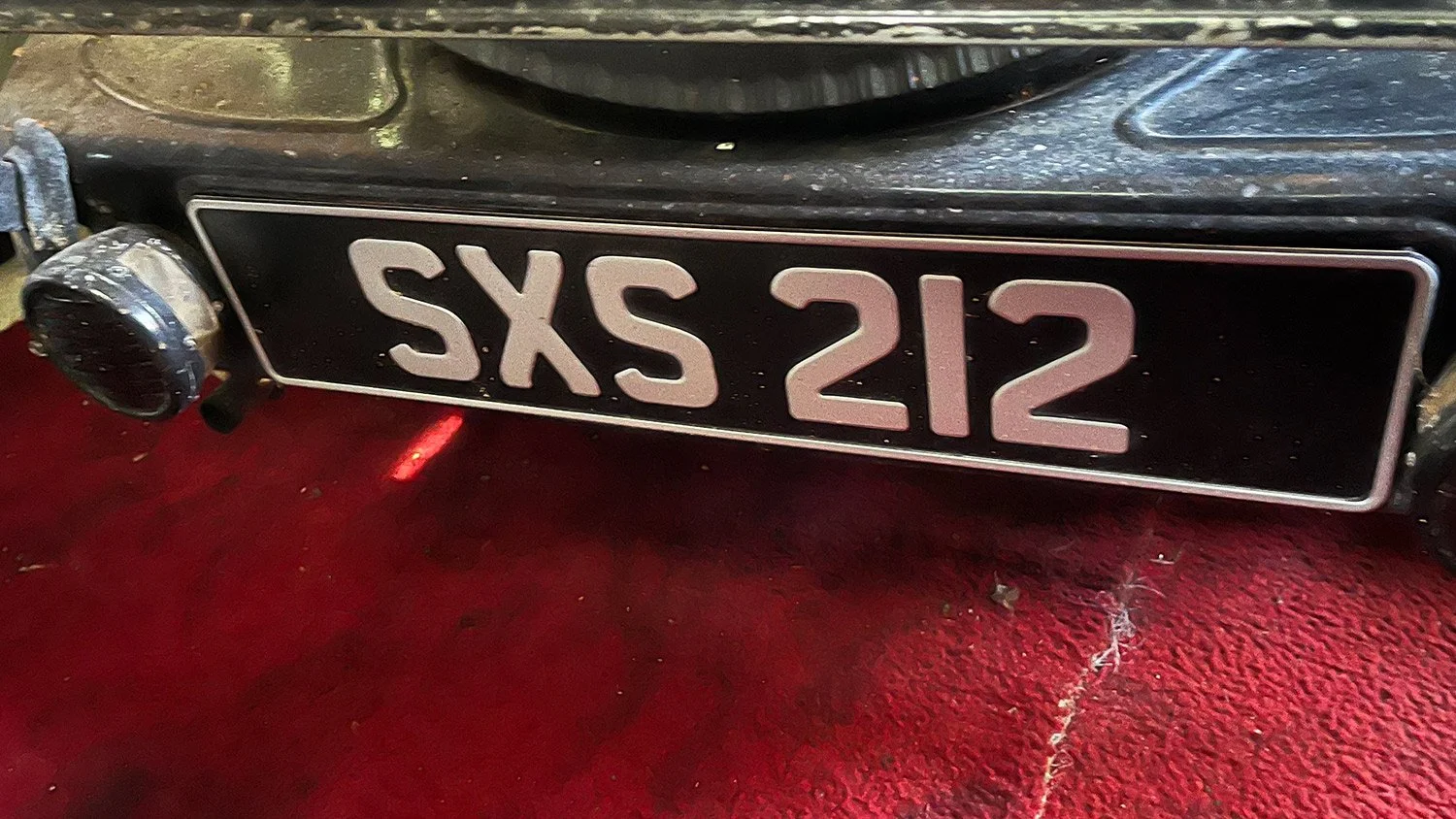 Close-up of old black vehicle license plate reading SXS 212 mounted on the front bumper, with a chrome bumper and red carpeted surface beneath.