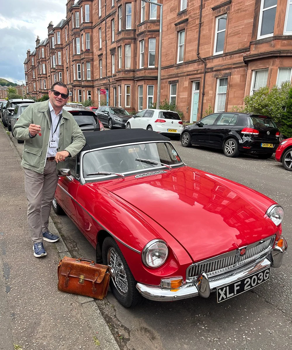 A man standing next to a red vintage MG convertible car with a black soft top, park on a city street, holding a set of keys, with a briefcase on the ground nearby.
