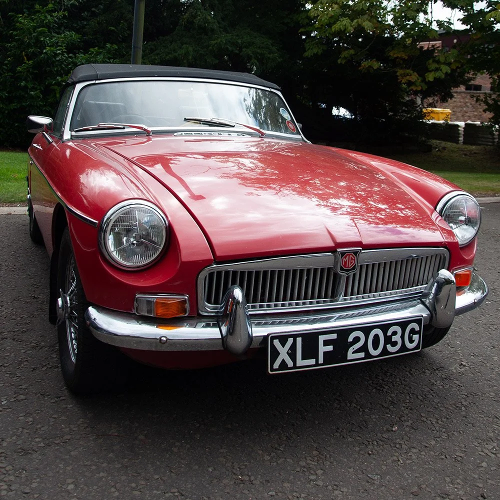 A red vintage MG convertible sports car parked on a driveway with trees and foliage in the background.