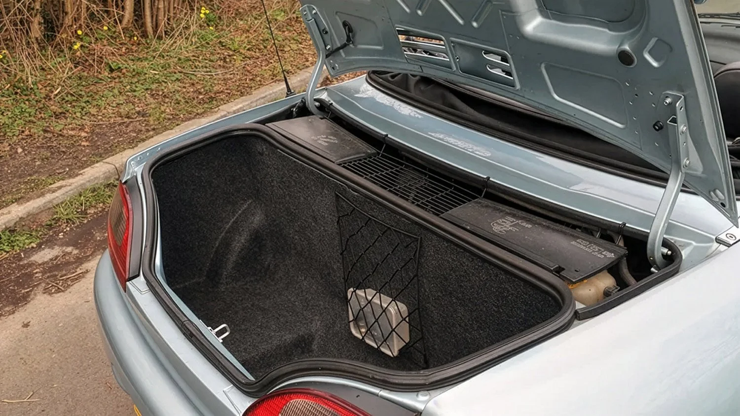 The open trunk of a silver car showing a black interior with a small netted compartment on the side and a large battery on the right.