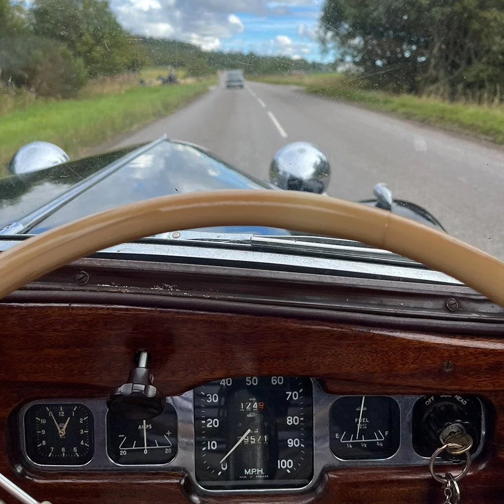 View from inside a vintage car looking at the dashboard and the road ahead, with trees and clouds in the sky.