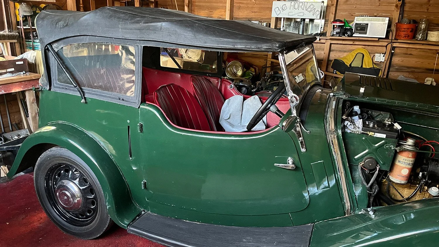 A vintage green open-top car with red seats and a black soft top, parked inside a wooden garage with shelves and tools visible in the background.