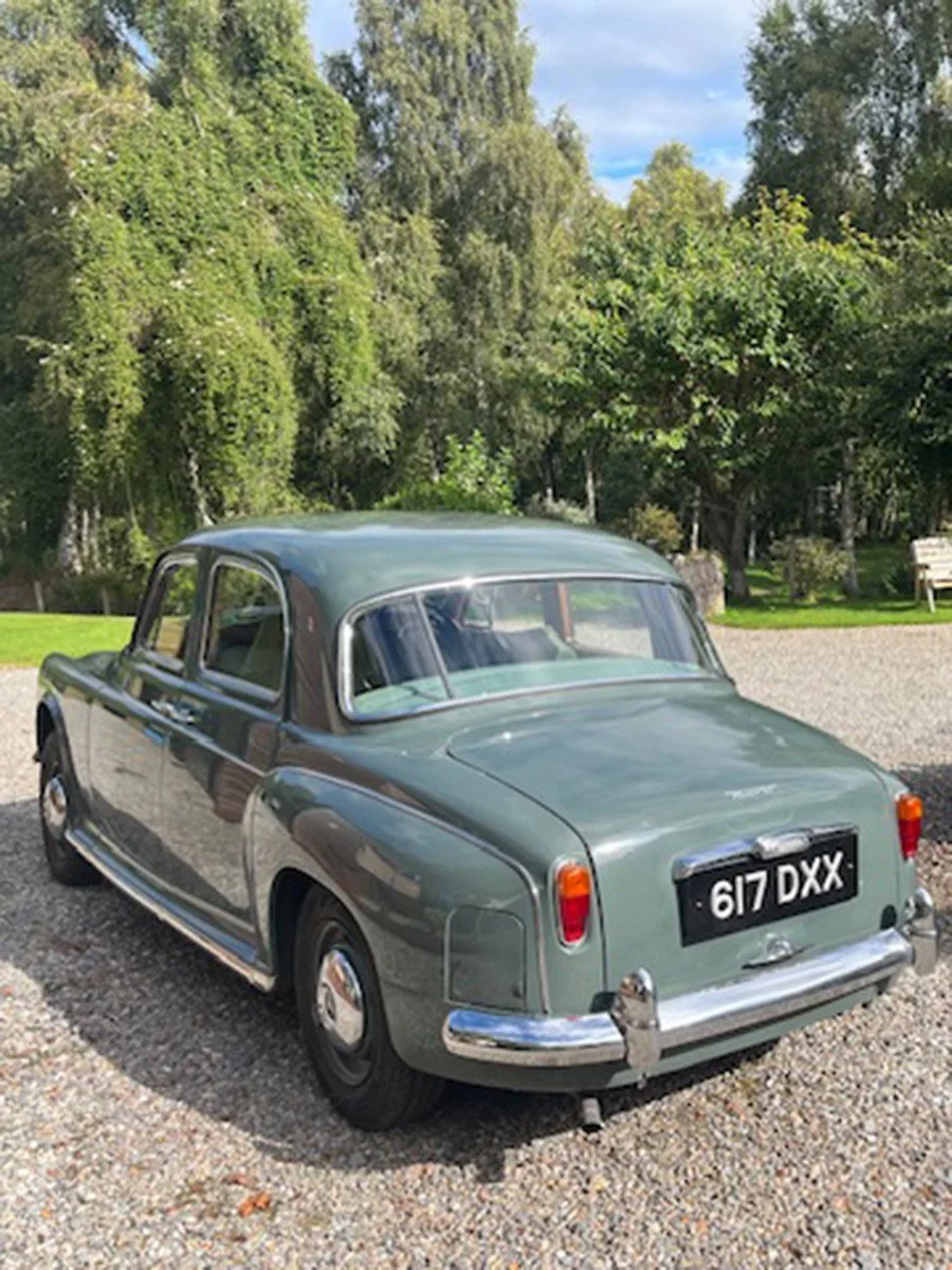 A vintage light green car parked on a gravel driveway with trees and green foliage in the background.