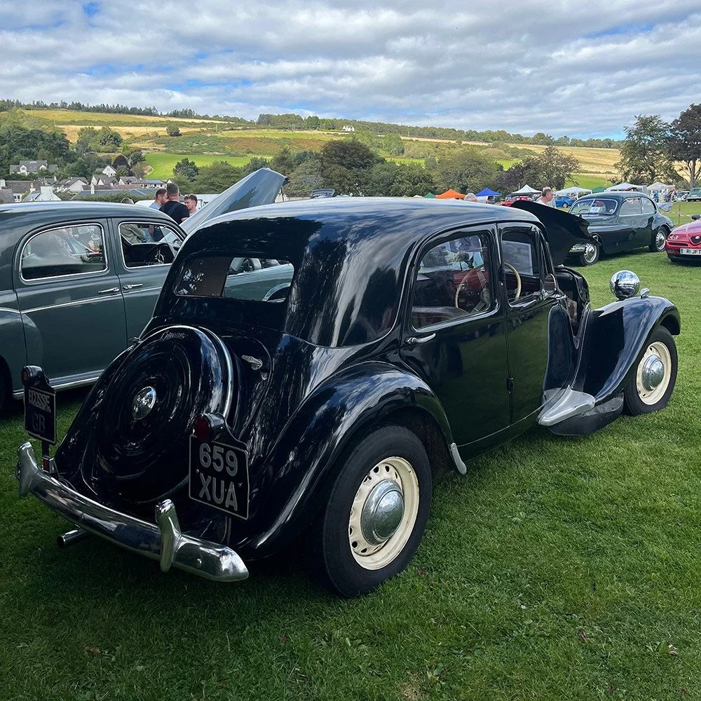 Black vintage car at a classic car show on a grassy field with other cars and tents in the background, and rolling hills under a partly cloudy sky.