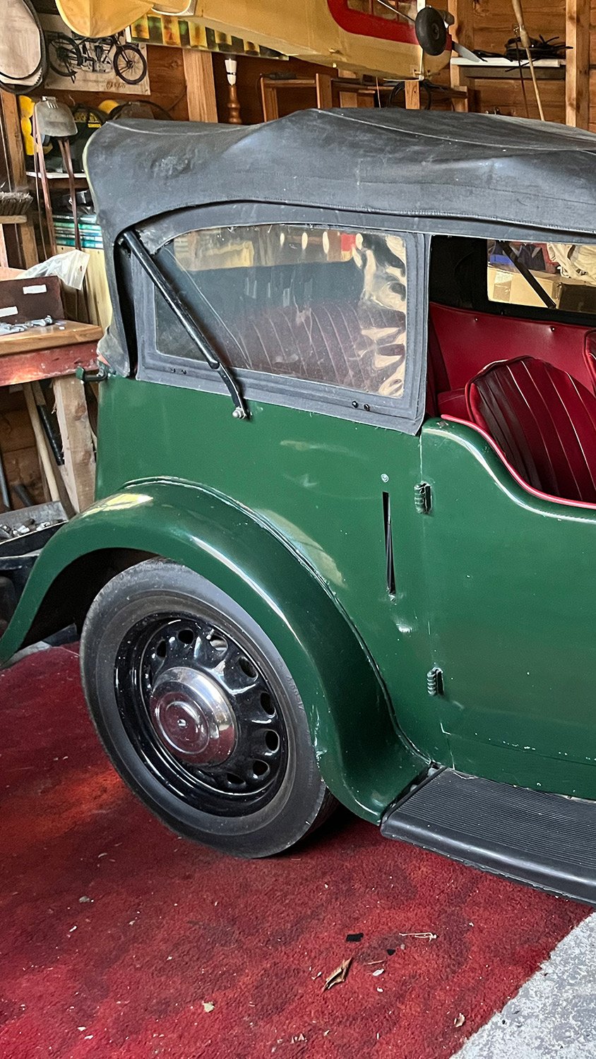 A vintage green car with a black soft top, red interior, and black wheel, parked inside a wooden garage.