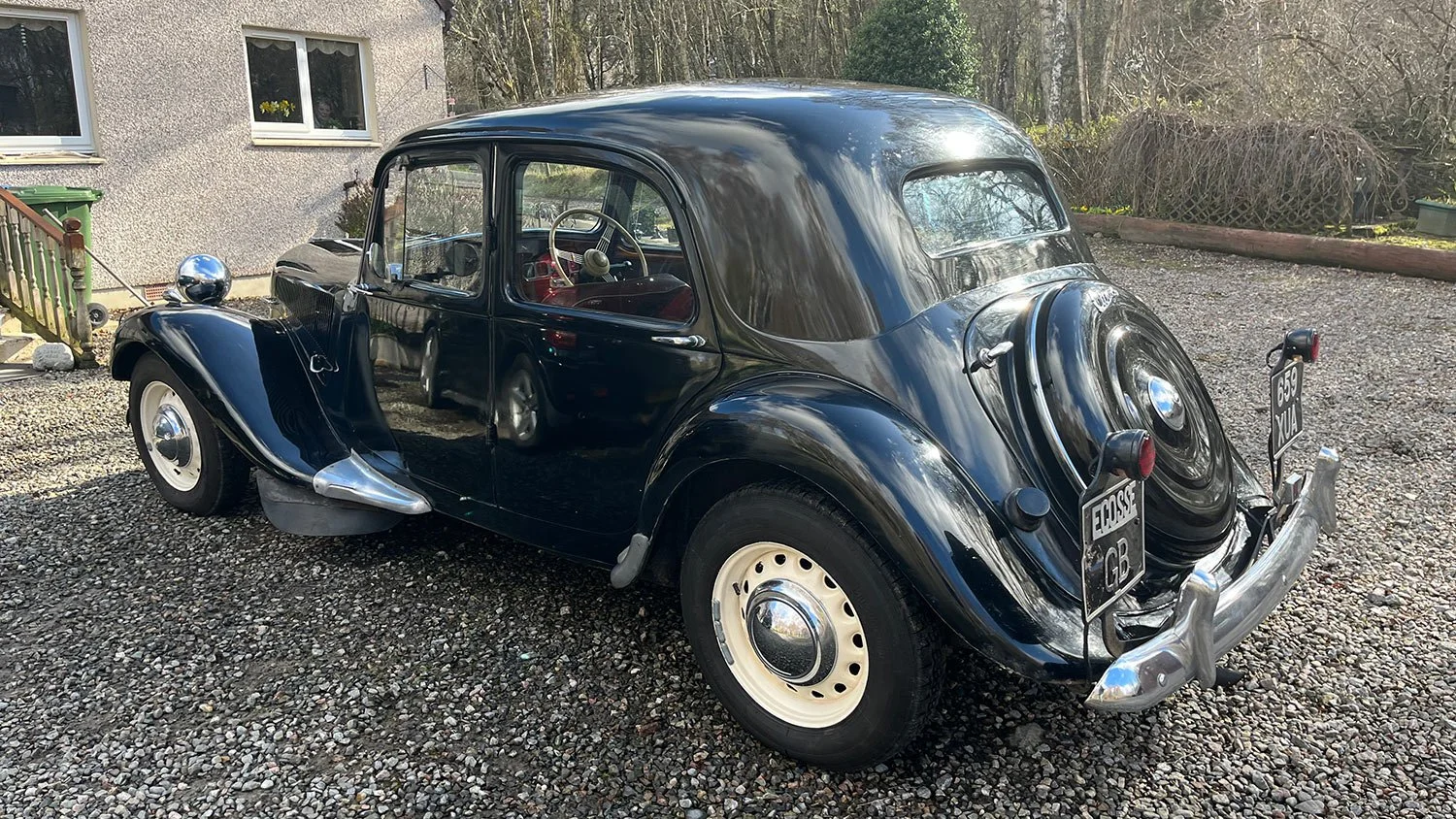 A vintage black car with white hubcaps parked on a gravel driveway next to a house with white window frames and a garden.