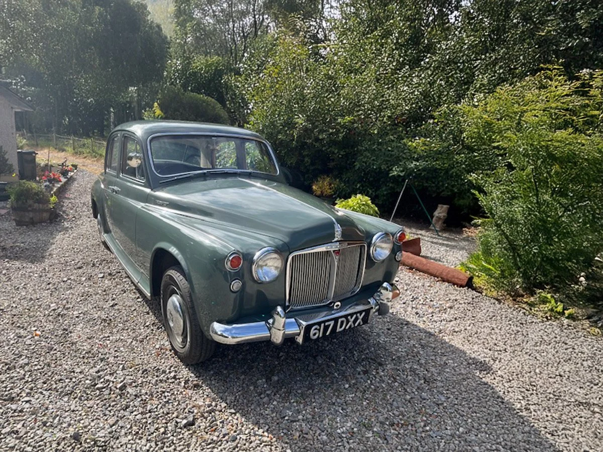 A vintage green car parked on a gravel driveway with greenery and plants in the background.