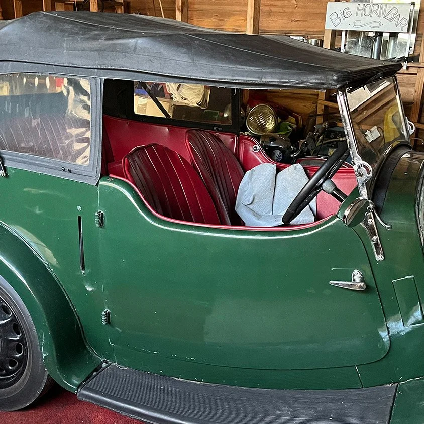 A vintage green car with a black soft top and red leather seats, parked inside a wooden garage with a sign that reads 'Big Horn Bar' in the background.