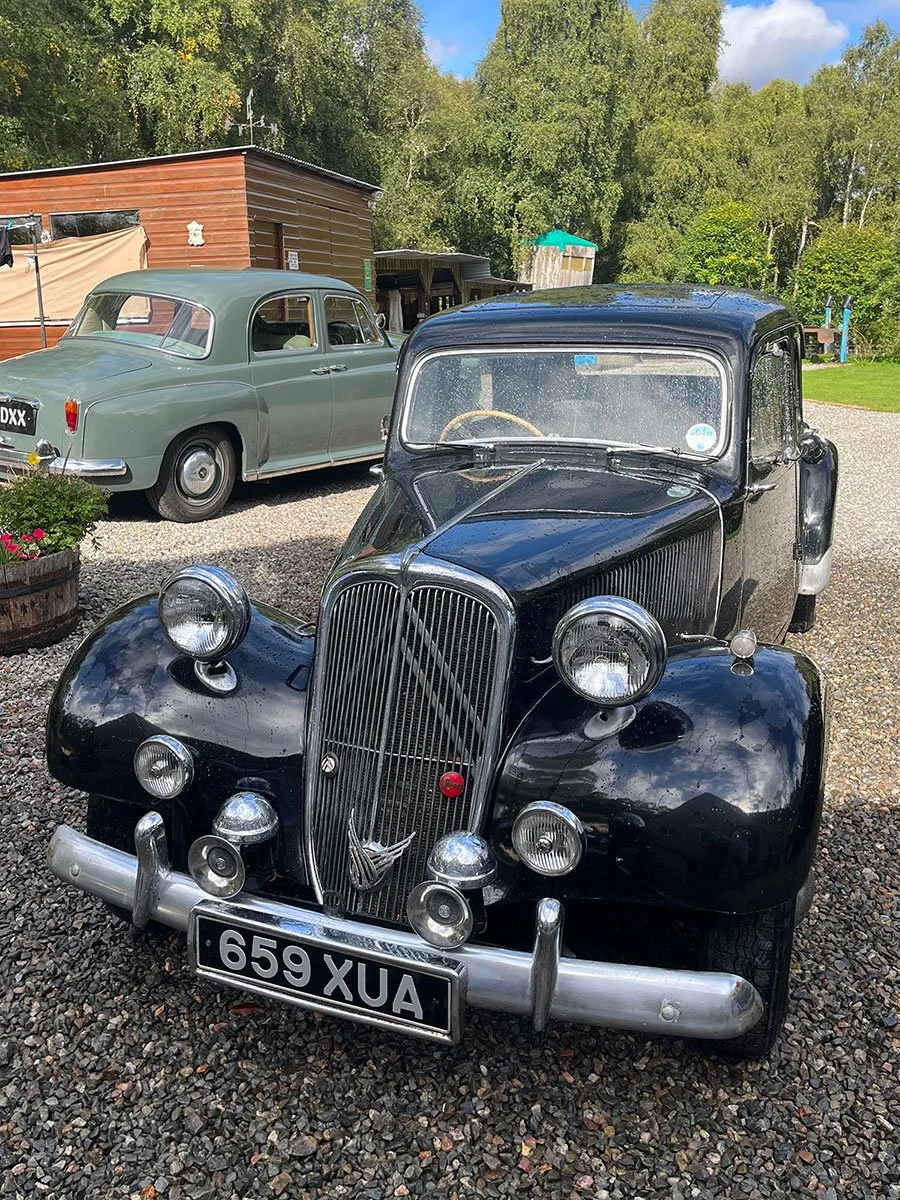 A vintage black car with a front grille, multiple round headlights, and a license plate reading '659 XUA', parked on a gravel surface. In the background, there is a green classic car parked beside a brown building, with trees and a partly cloudy sky 