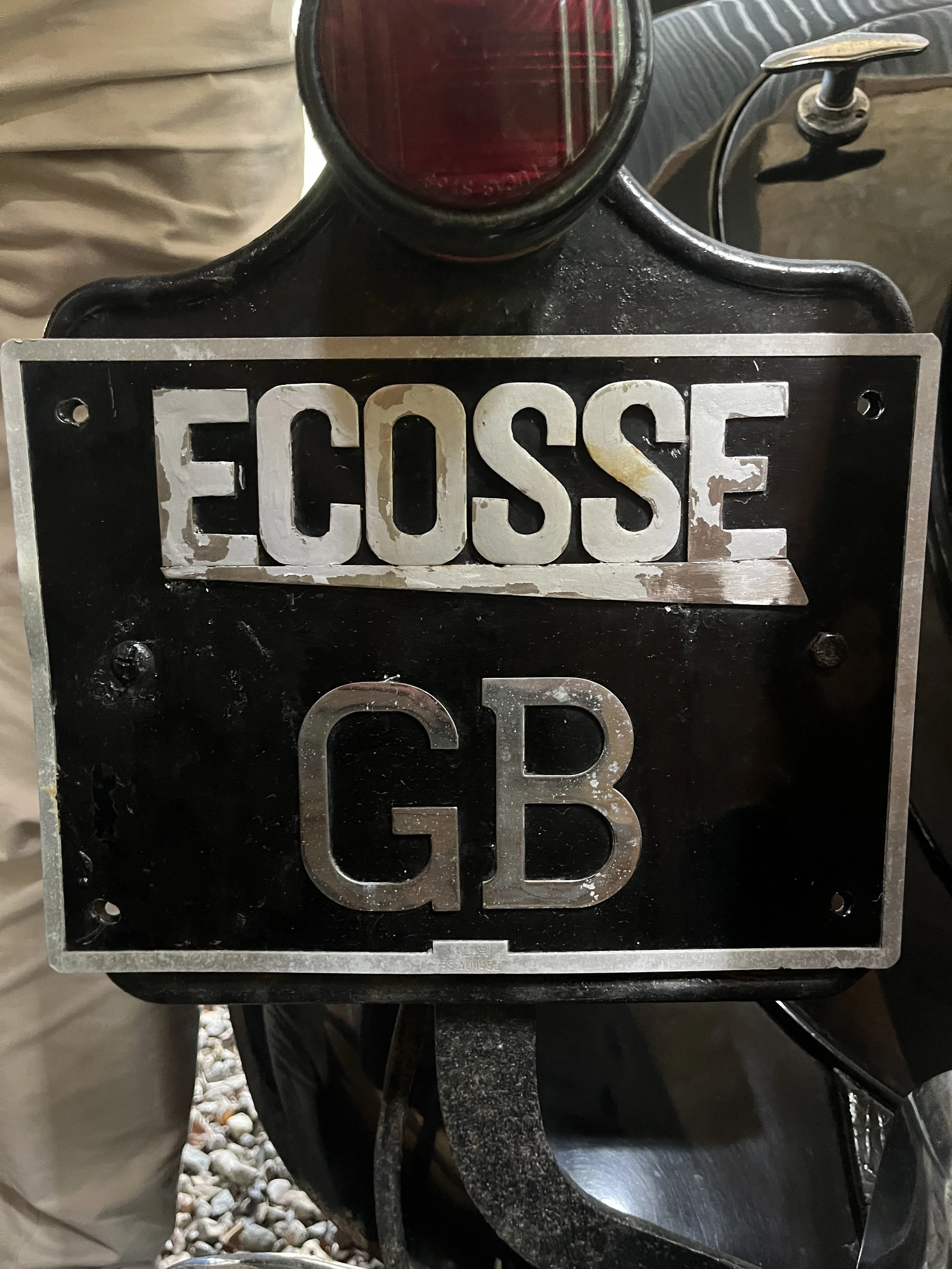 A vintage metal sign with the words 'ECOSSE GB' mounted on a black vehicle, possibly a motorcycle or bicycle, with a red rear light visible on top.