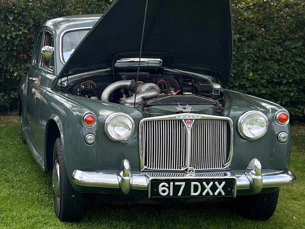 A vintage Rover car with its hood open, parked on grass with bushes in the background.